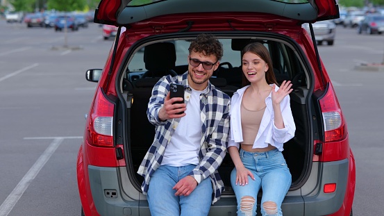 Romantic couple sitting on the trunk car and posing at smartphone camera, waving hands and making selfie. Tansportation, technology, trip, travel and lifestyle concept. Slow motion