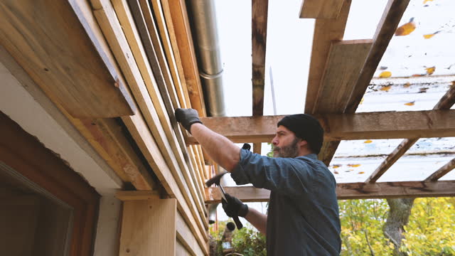 bearded man nailing wooden weatherboards at facade