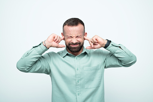 Mid adult men wearing green shirt covering ears with fingers and with eyes closed. Studio shot, white background.