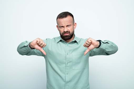 Mid adult men wearing green shirt standing with thumbs down and looking at camera. Studio shot, white background.