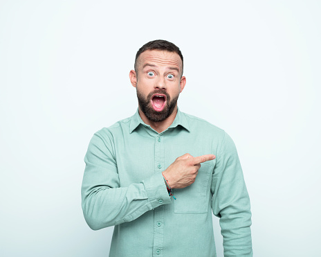 Surprised mid adult men wearing green shirt pointing with index finger at copy space, staring at camera with mouth open. Studio shot, white background.