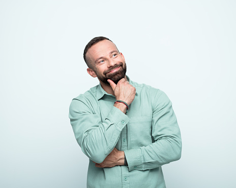 Pleased mid adult men wearing green shirt standing with hand on chin, looking up and smiling. Studio shot, white background.