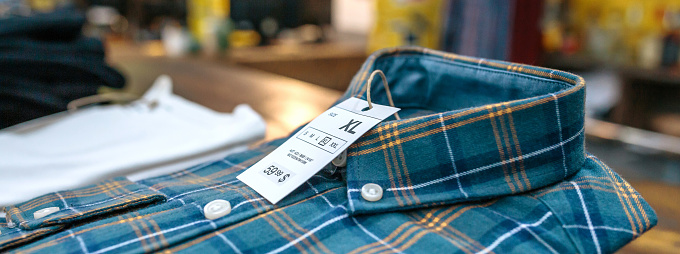 Detail of label with price and size over a blue plaid shirt on industrial style store. Banner of organized apparel stacks over counter ready to sell on vintage clothes shop.