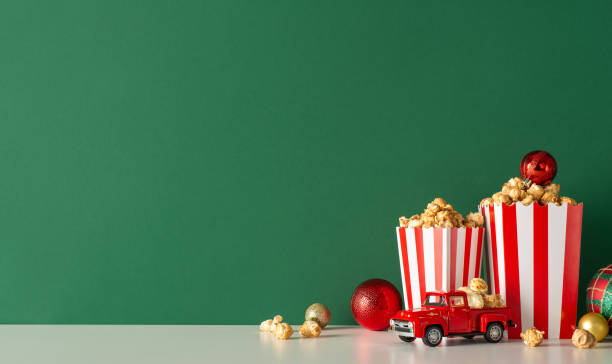istock Create festive atmosphere for Noel with home popcorn delivery idea. Side view of tabletop, featuring charming red car, delectable popcorn, decorative balls. Green wall backdrop for movie ads or promo 1778455513
