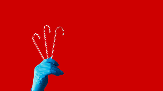 Candy canes. Pandemic holiday. Human hand in blue protective glove holding peppermint sweetmeats isolated on red copy space background.