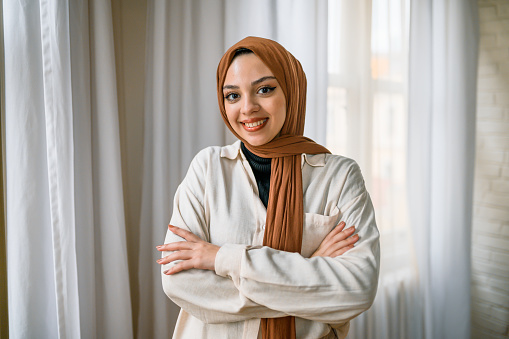 Cheerful young muslim woman wearing hijab crossing arms and smiling at camera in room