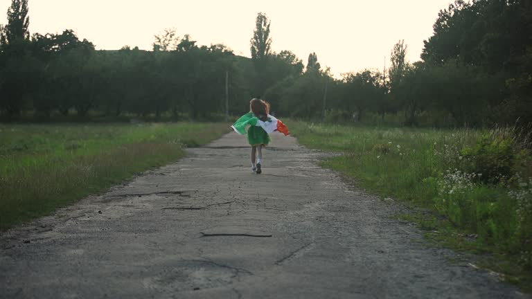 Little girl with red hair running outdoors covered with Irish flag on Saint Patrick's Day