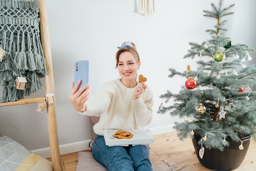 Happy Young Woman in cozy sweater taking selfie photo while relaxing on floor cushion near potted christmas tree in modern Scandi interior home. Cozy winter holiday. Content creating. Selective focus