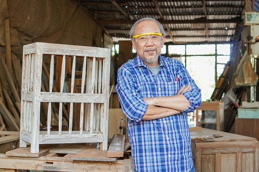 Portrait of Asian senior male carpenter in arms crossed pose in the workshop with wood boards background. copy space