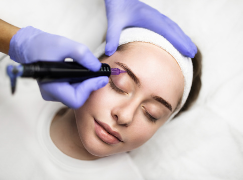 Beautician hand doing eyebrow permanent makeup on young attractive female face, professional pmu artist in gloves making microblading tattoo to brows of beautiful woman, using machine pen, closeup