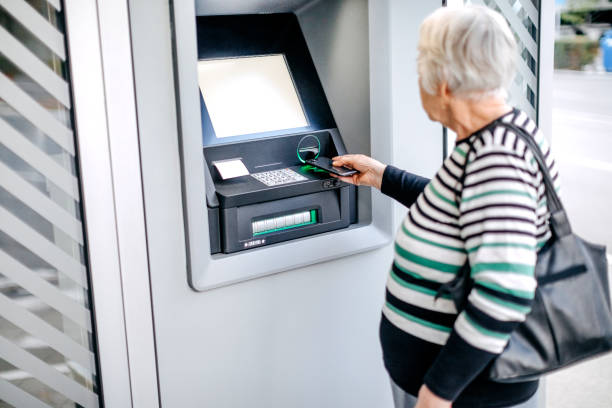 istock Old woman picking money using smart phone in front of ATM 1778643172
