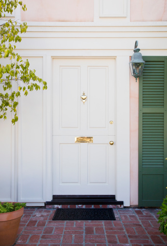 Vertical shot of a white front door of a home with green shutters, brick flooring and brass accents.