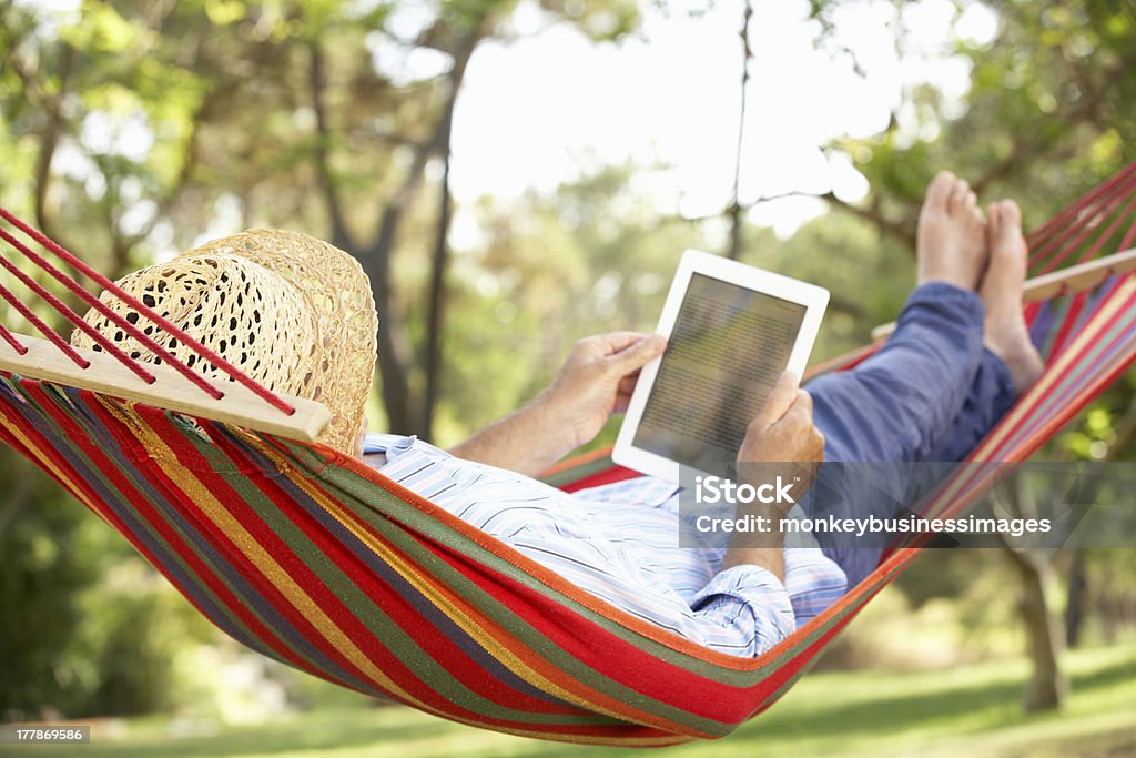 Man wearing hat relaxing in hammock with e-book Senior Man Relaxing In Hammock With E-Book Lying Down Wearing Straw Hat Hammock Stock Photo