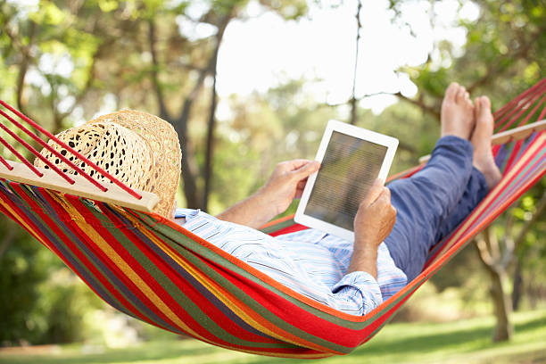 Man wearing hat relaxing in hammock with e-book Senior Man Relaxing In Hammock With E-Book Lying Down Wearing Straw Hat hammock stock pictures, royalty-free photos & images