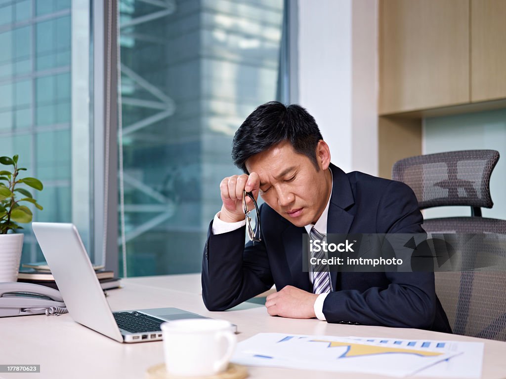 Stressed businessman rubbing head looking at charts "asian businessman sitting in office, looking tired. click for more: abusiness peopleai" Asian and Indian Ethnicities Stock Photo