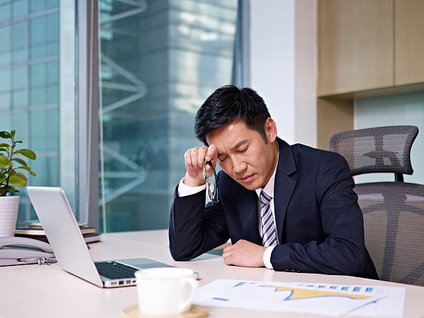 Stressed businessman rubbing head looking at charts "asian businessman sitting in office, looking tired. click for more: abusiness peopleai" face down stock pictures, royalty-free photos & images