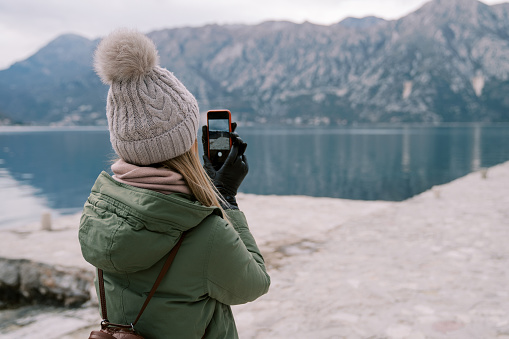 Woman stands on the seashore and takes pictures of the mountains on a smartphone. Back view. High quality photo