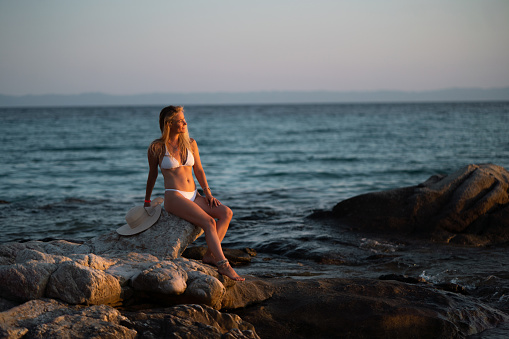 Happy woman in swimsuit sitting on the rock at the beach and looking at view