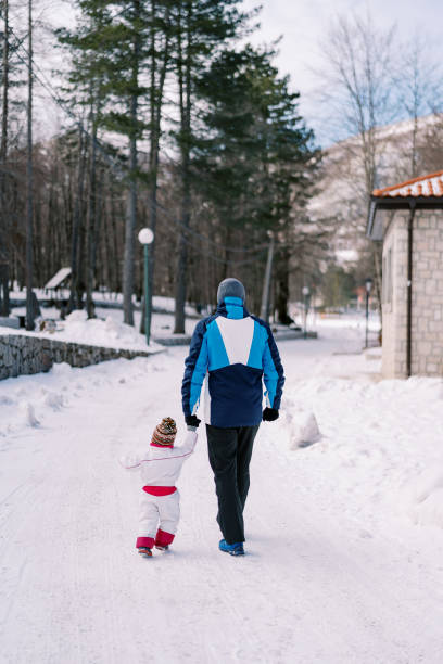 dad and a little girl walk along a snowy road past the house, holding hands. back view - 5105 imagens e fotografias de stock
