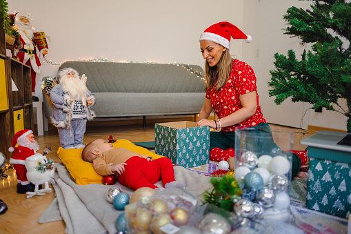 A mother busily wraps Christmas gifts while her baby girl peacefully lays on the floor nearby.