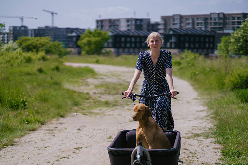 Embrace urban nature with a woman pedaling her cargo bike, accompanied by her dog, through a sunlit city park. A perfect image of city living and pet-friendly activities.