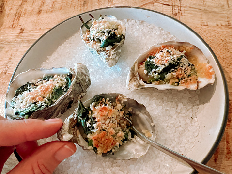 Horizontal high angle closeup photo of four freshly shucked Australian oysters in the half shell with Bechamel sauce, spinach and grilled breadcrumbs served on a bed of rock salt on a ceramic plate on a wooden table in a fine dining restaurant. A woman’s fingers are holding one of the oyster shells. Byron Bay, NSW.