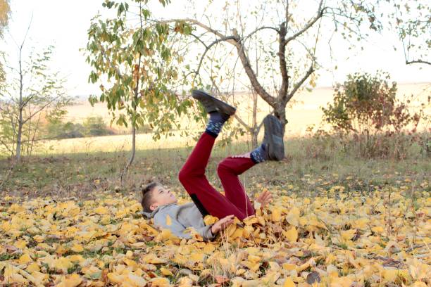 A cheerful toddler sits barefoot on a leaf-strewn path, wearing a red beanie and blue outfit. This warm autumn scene captures playful innocence, nature, and candid joy.