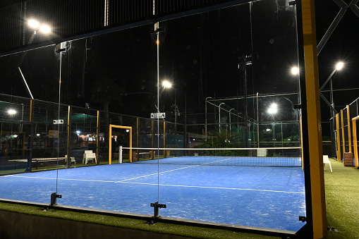 Night view on enclosed blue court for padel with construction created by mesh and the glass back walls. Padel is a racket sport played in doubles and is similar to tennis with rules of scoring.