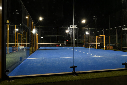 An enclosed blue court for padel in the night with construction created by mesh and the glass back walls. Padel is a racket sport played in doubles and is similar to tennis with rules of scoring.