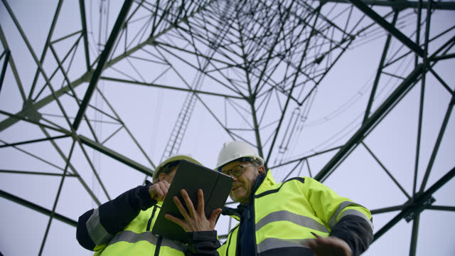SLO MO Electrical Engineers Looking Up At Electricity Pylon And Using Digital Tablet At Power Station
