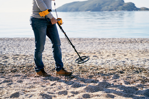 Beach cop with a metal detector walks along the beach, sweeping it over the pebbles. Cropped. High quality photo