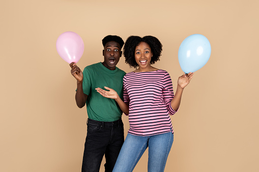 Cheerful surprised young black man and lady with big belly enjoy pregnancy and expect baby, holding inflatable balloons, isolated on beige background, studio. Parents and family and gender party