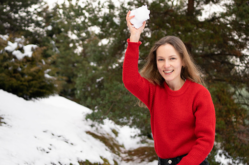 A young woman in a red sweater is preparing to throw a snowball for a snowball game against a background of Christmas trees. The concept of winter holidays, having fun and outdoor activities