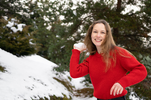 A young attractive woman in a red sweater is preparing to throw a white snowball while playing a snowball fight outdoor. Concept of winter entertainment and activities