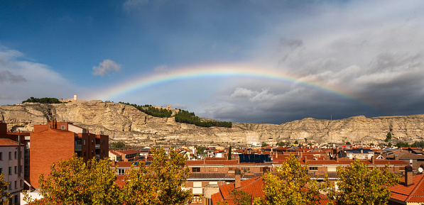 Capture the breathtaking beauty of a complete rainbow arching over the charming city of Calatayud, with its ancient castle in the foreground, set against a dramatic backdrop of strong winds and turbulent weather. This mesmerizing image is a must-have for stock agencies, showcasing nature's majesty in its most dramatic form.