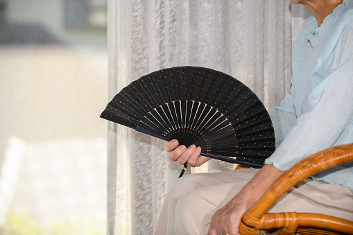 A senior woman sits on a rattan chair while cooling herself with a fan in the hot summer window.