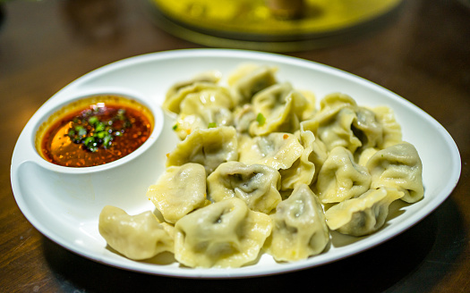 Stock photo showing an evening meal of steamed dumplings (Momos), filled with mixed vegetables and pork drizzled with chilli oil and garnished with chopped spring onion and coriander leaves on plate with red and orange spicy dipping sauce.