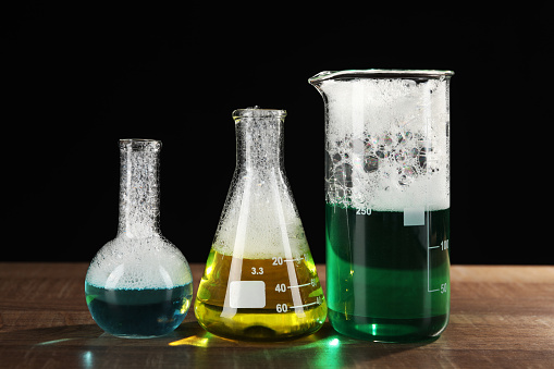 Laboratory glassware with colorful liquids on wooden table against black background. Chemical reaction