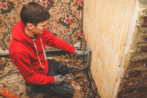 A young Caucasian man in a red sweatshirt squats and removes retro wallpaper from the wall using a steam generator in an old abandoned house, close-up top view. Concept for home renovation, construction work, home.