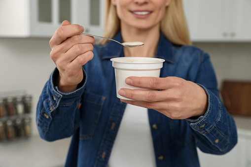 Woman holding plastic cup and spoon with tasty yogurt in kitchen, closeup