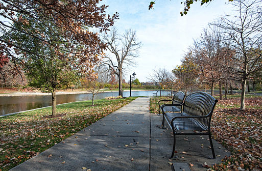 Urban park with black wrought iron benches and sidewalk leading to pond in late fall.