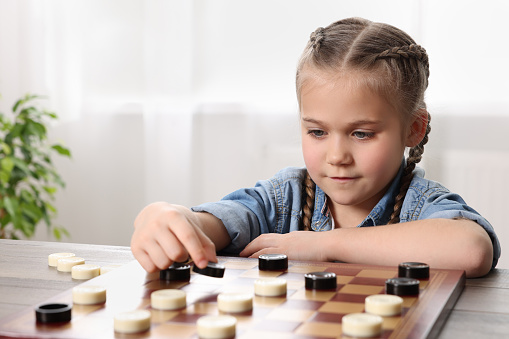 Playing checkers. Little girl thinking about next move at table in room