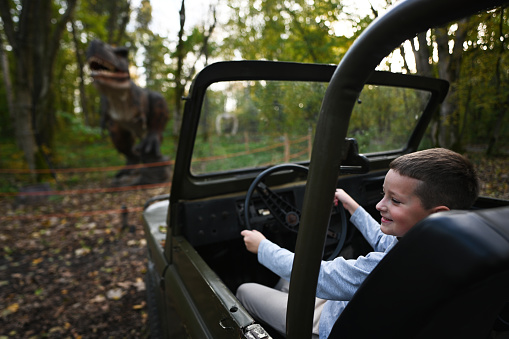Boy driving a convertible car with dinosaurs in the background, selective focus.