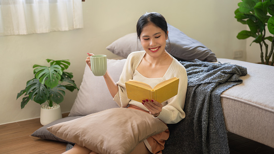 A happy and charming young Asian woman is sitting on the floor beside her bed, enjoying her morning coffee and reading a book in her bedroom. Leisure, domestic life