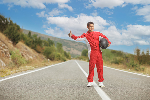 Full length shot of a racer in a red suit holding a helmet and hitchhiking on an open road