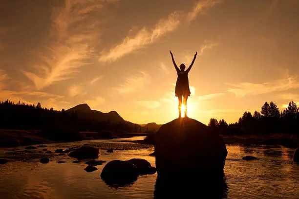 A silhouette of a young woman in Sun Salutation yoga pose series on the Tuolumne River in Yosemite at sunset.