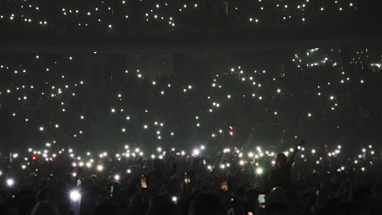 Rear view of large group of people enjoying a concert performance. There are many hands applauding and taping the show. Multi colored lasers and spot lights firing from the stage.\nSilhouettes have been significantly liquified.