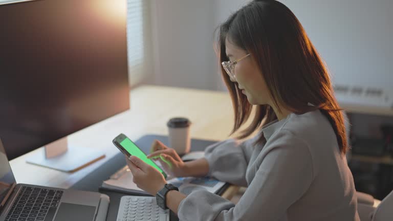 Businesswoman using smartphone green screen after working with laptop on table, woman using mobile phone search or social media or shopping online or stock or crypto currency. Smart phone conversation conference.