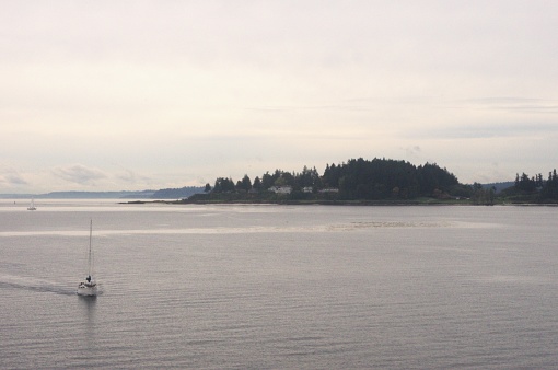 Sailboat with sail packed away, traveling via motor in a bay with almost no breeze, with Mt Rainer partially hidden by the clouds in the distance. Taken on the ferry between Seattle and Bainbridge Island, which is traveling in the Puget Sound in Washington, USA.