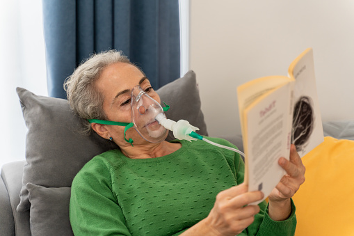 Senior woman with nebulizer inhaler face mask reads book at home